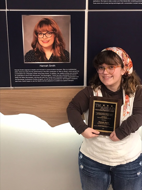 A student holds a plaque with her name on it and stands in front of a picture of herself as a member of the 1911 society. 
